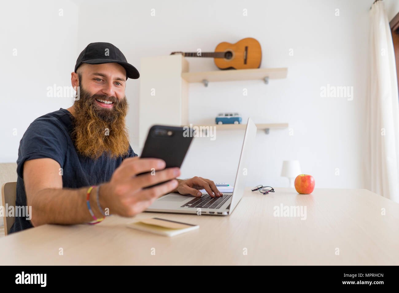 Portrait de jeune homme barbu assis à un bureau avec ordinateur portable looking at cell phone Banque D'Images