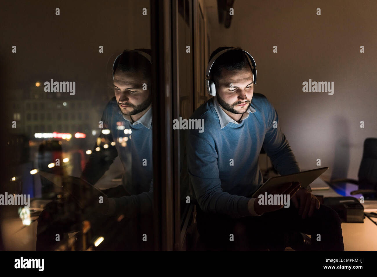 Businessman sitting on window sill dans office, la nuit, un casque et tablette Banque D'Images