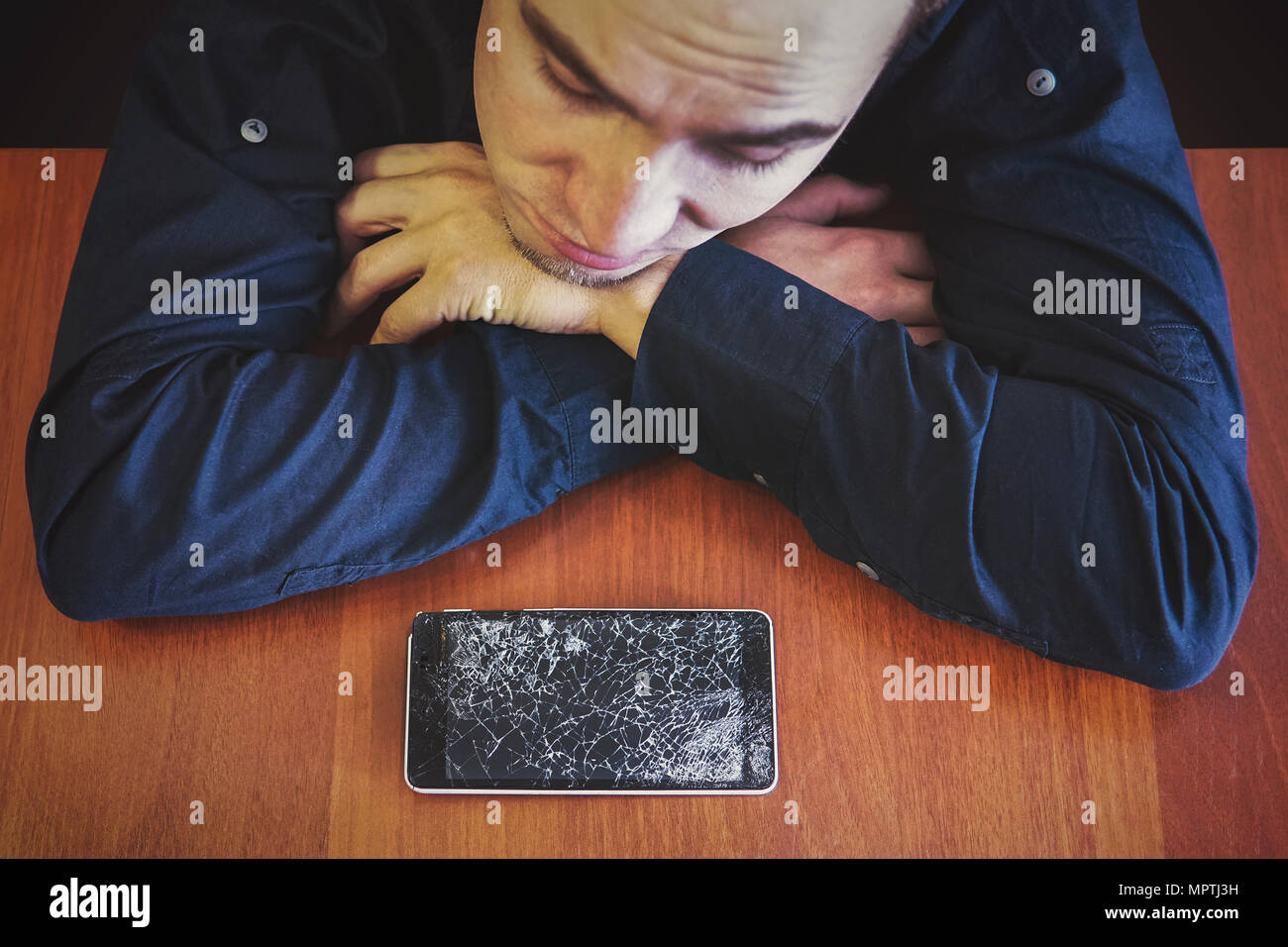 Un homme regarde son téléphone cassé posé sur une table en bois avec un regard triste. Guy s'est fâché au sujet de casser son téléphone. Shattered phone afficher Banque D'Images