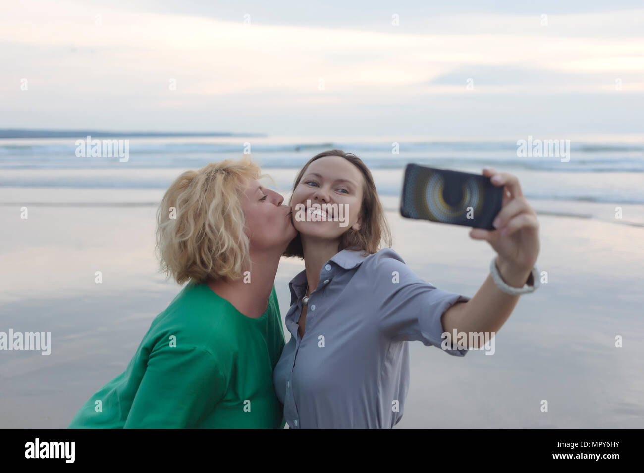 Cheerful woman with smart phone selfies lors d'être embrassé par ami à la plage pendant le coucher du soleil Banque D'Images