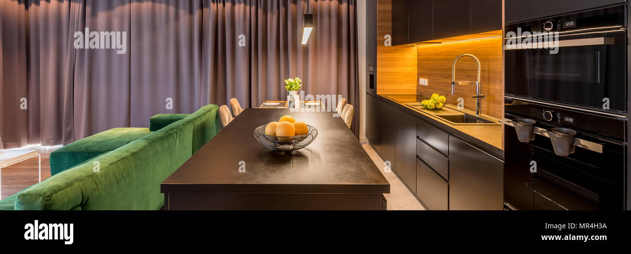 Panorama d'un espace ouvert salon intérieur avec un canapé vert à côté d'une cuisine moderne, d'un four et d'island avec un bol de fruits Banque D'Images