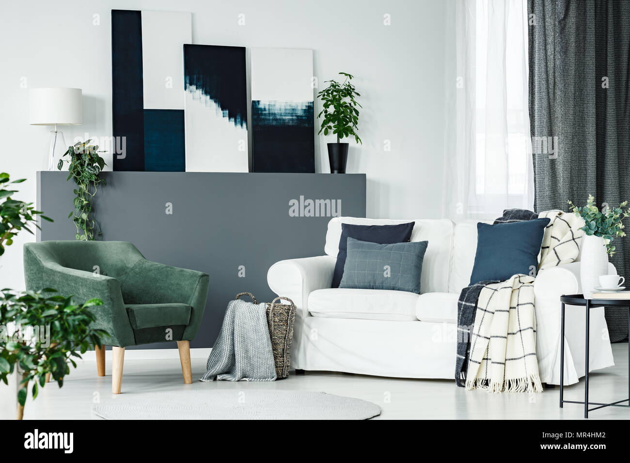 Couverture grise dans panier en osier placé entre vert fauteuil et canapé blanc avec des oreillers en salon lumineux avec des peintures d'intérieur et fenêtre Banque D'Images