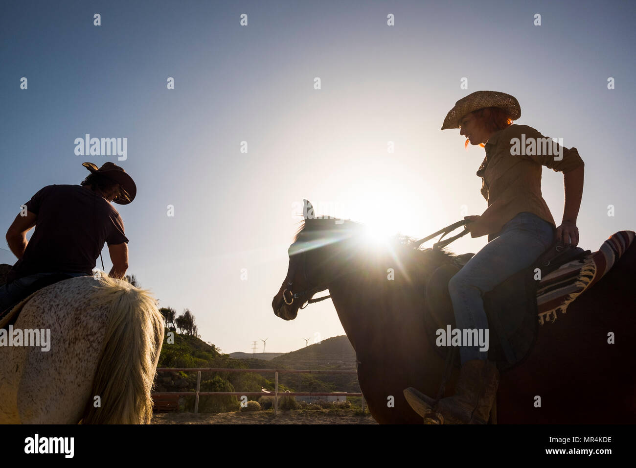 Couple de cow-boys modernes l'homme et la femme monter deux chevaux piscine avec sunflare et rétroéclairage. montagne et moulin à vent en arrière-plan. beau jeune sur vacat Banque D'Images