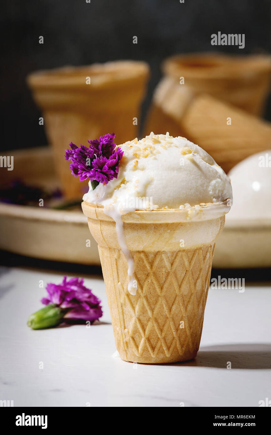 La crème glacée à la vanille en petits waffle tasse servi avec des fleurs comestibles et cuillère en métal dans une plaque en céramique sur une table de cuisine en marbre blanc. Banque D'Images