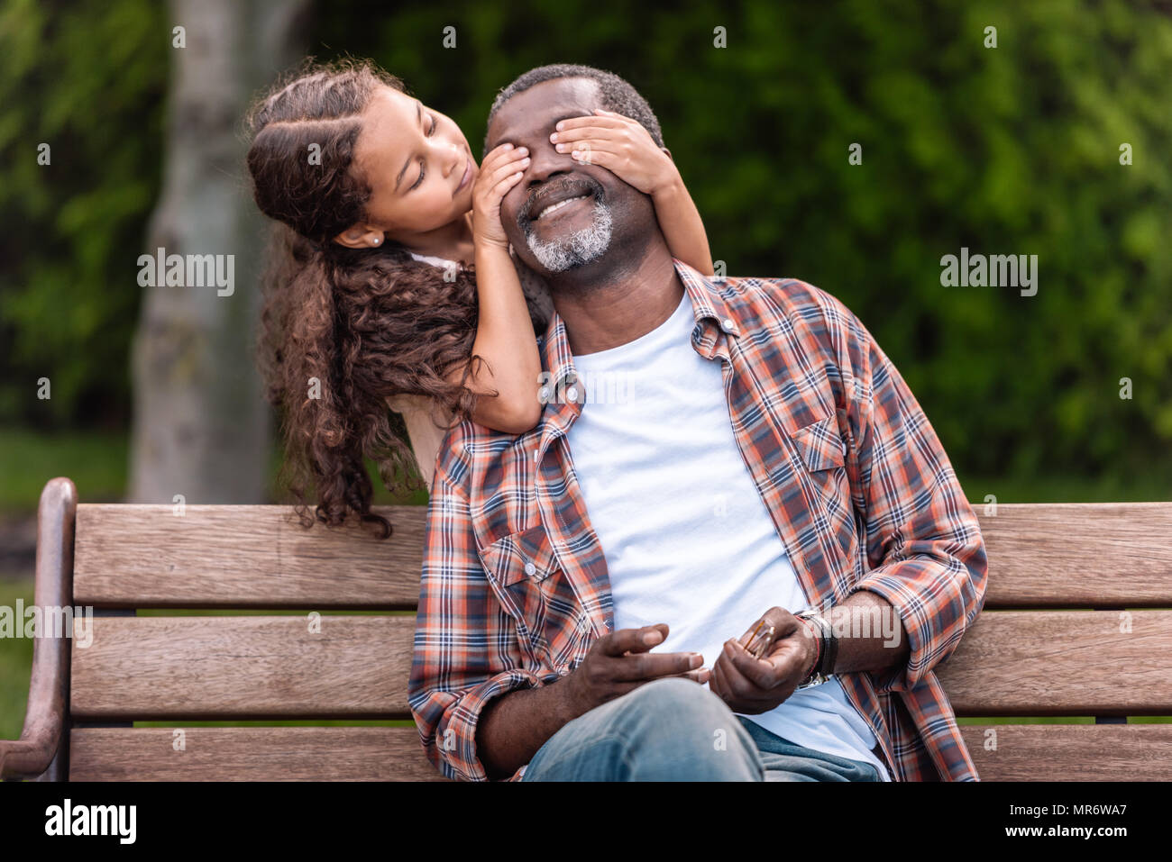 Smiling african american girl fermer les yeux de son grand-père assis sur un banc dans le parc Banque D'Images