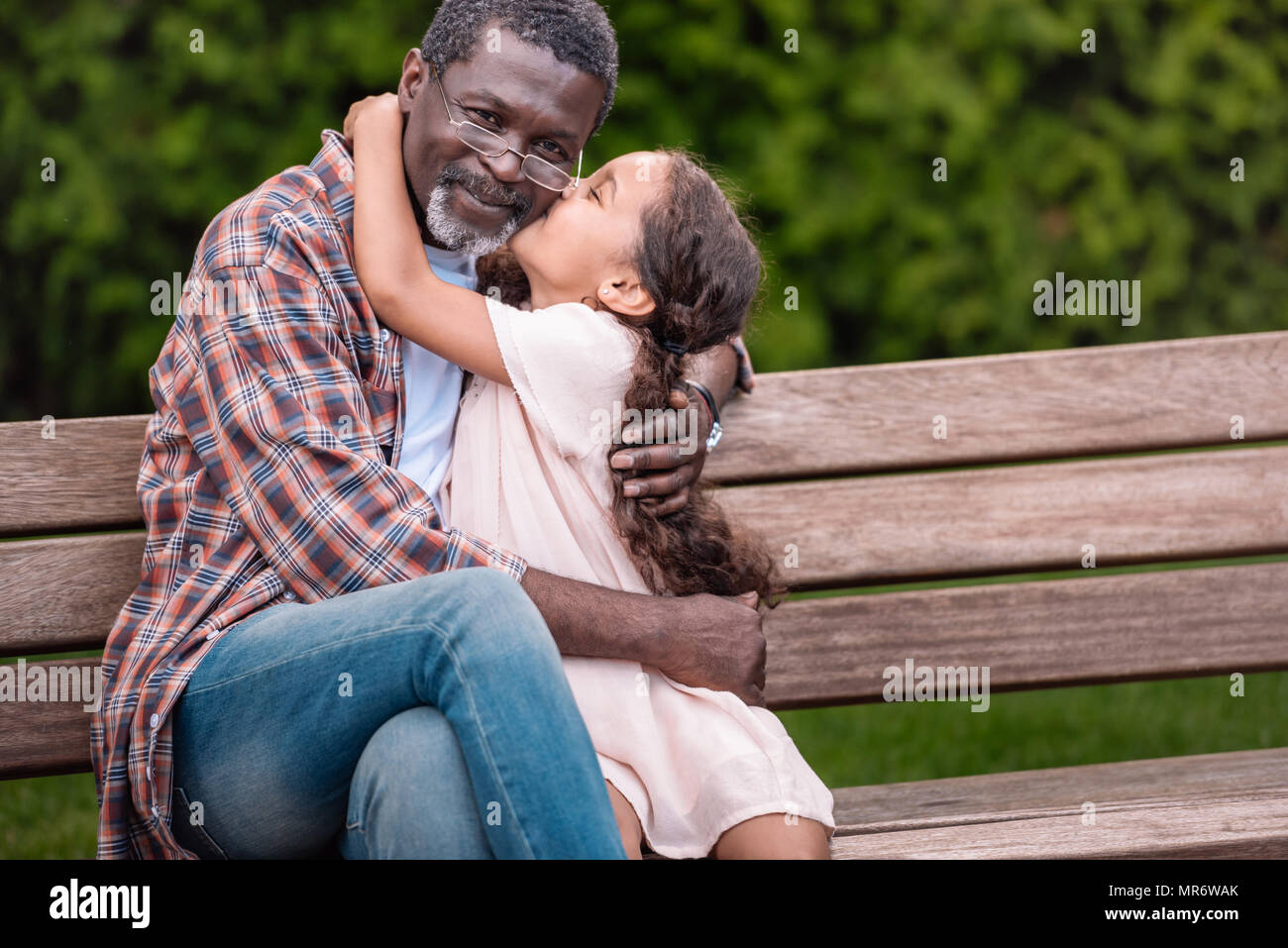 Adorable african american girl hugging et embrasser son grand-père alors qu'il était assis sur un banc dans le parc Banque D'Images