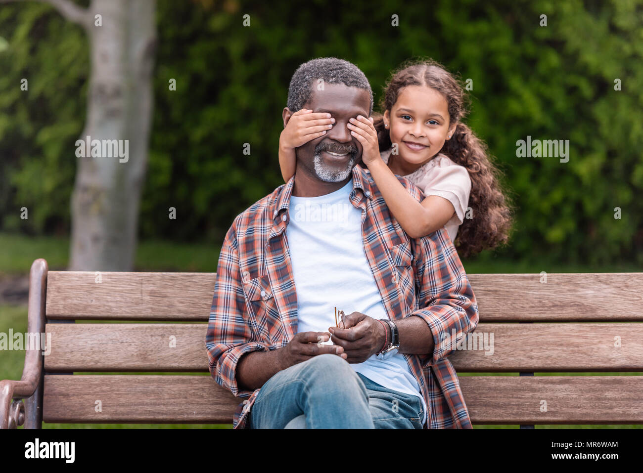 Smiling african american girl fermer les yeux de son grand-père assis sur un banc dans le parc Banque D'Images