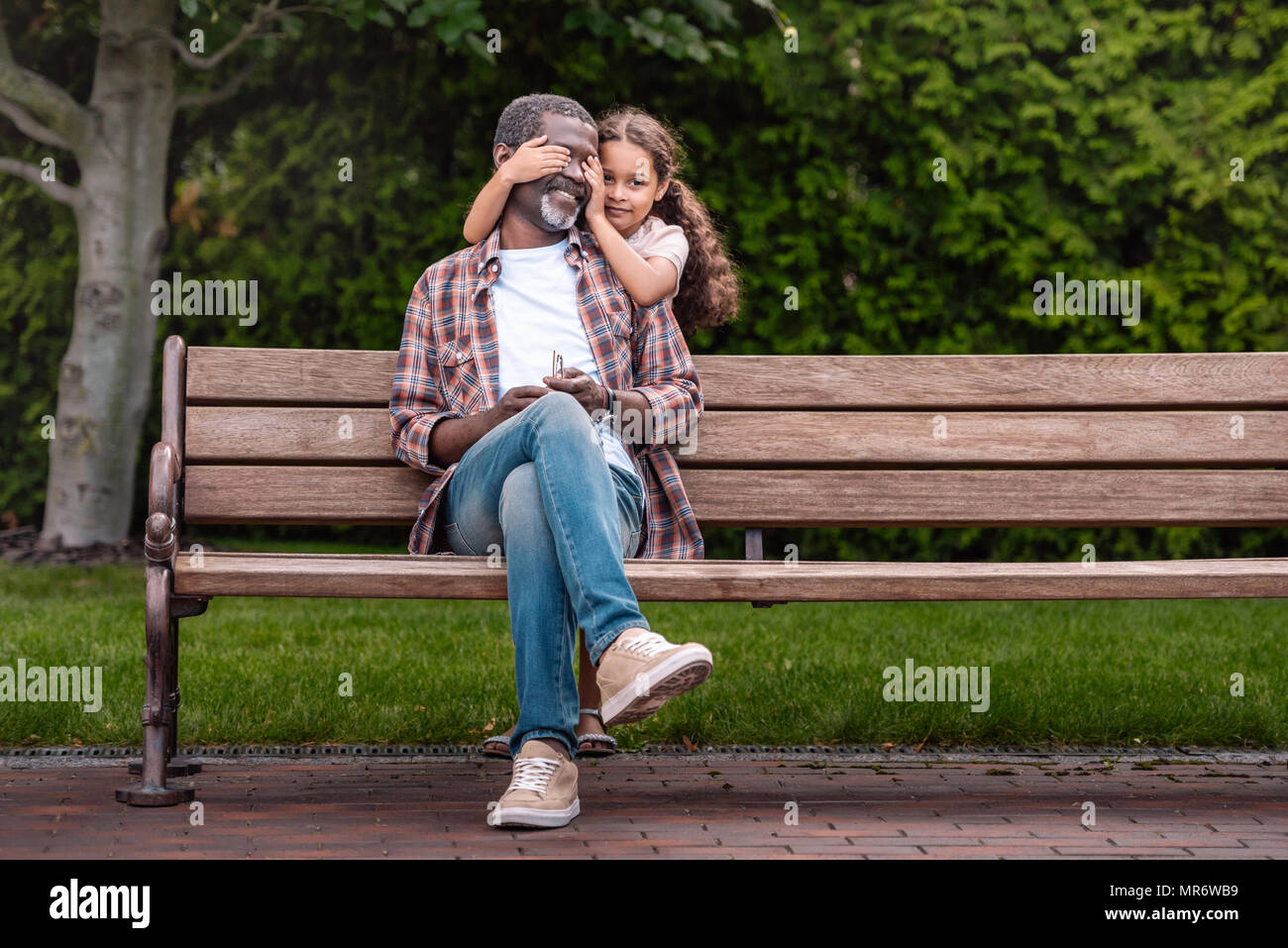 Cute african american girl fermer les yeux de son grand-père assis sur un banc dans le parc Banque D'Images