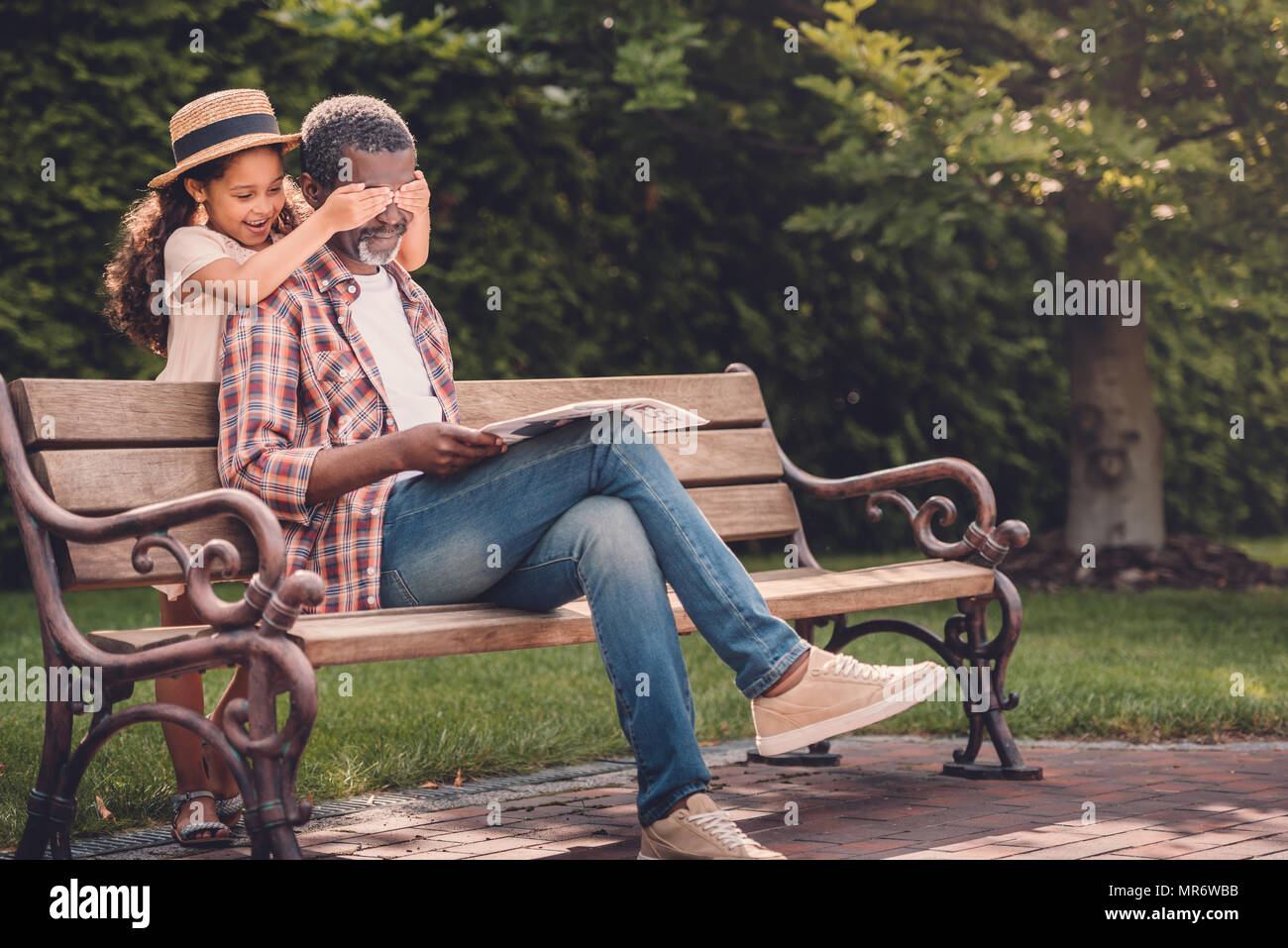 Smiling african american girl fermer les yeux de son grand-père assis sur un banc dans le parc Banque D'Images