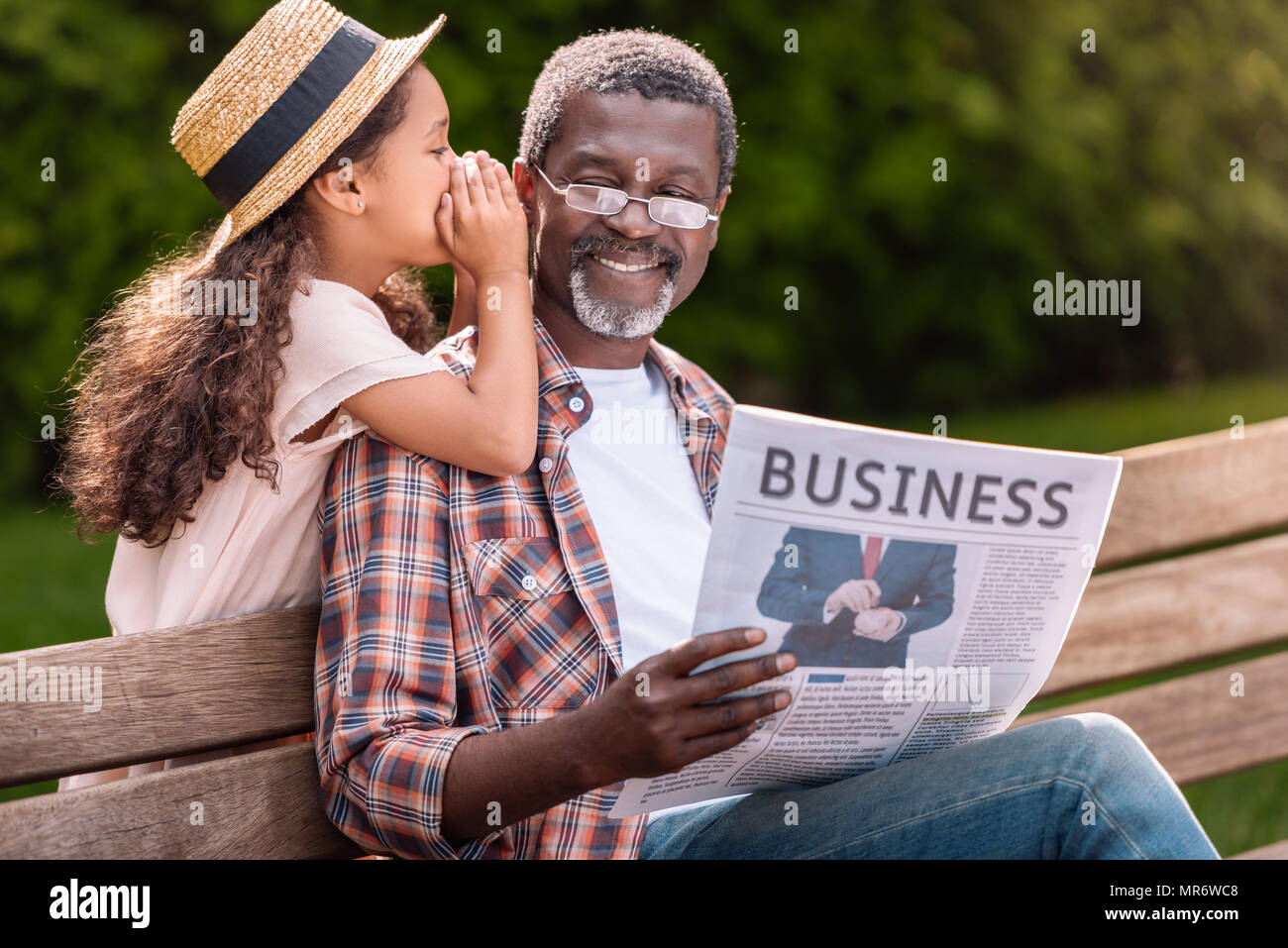 African American girl whispering à son grand-père alors qu'il lit le journal d'affaires sur l'établi Banque D'Images