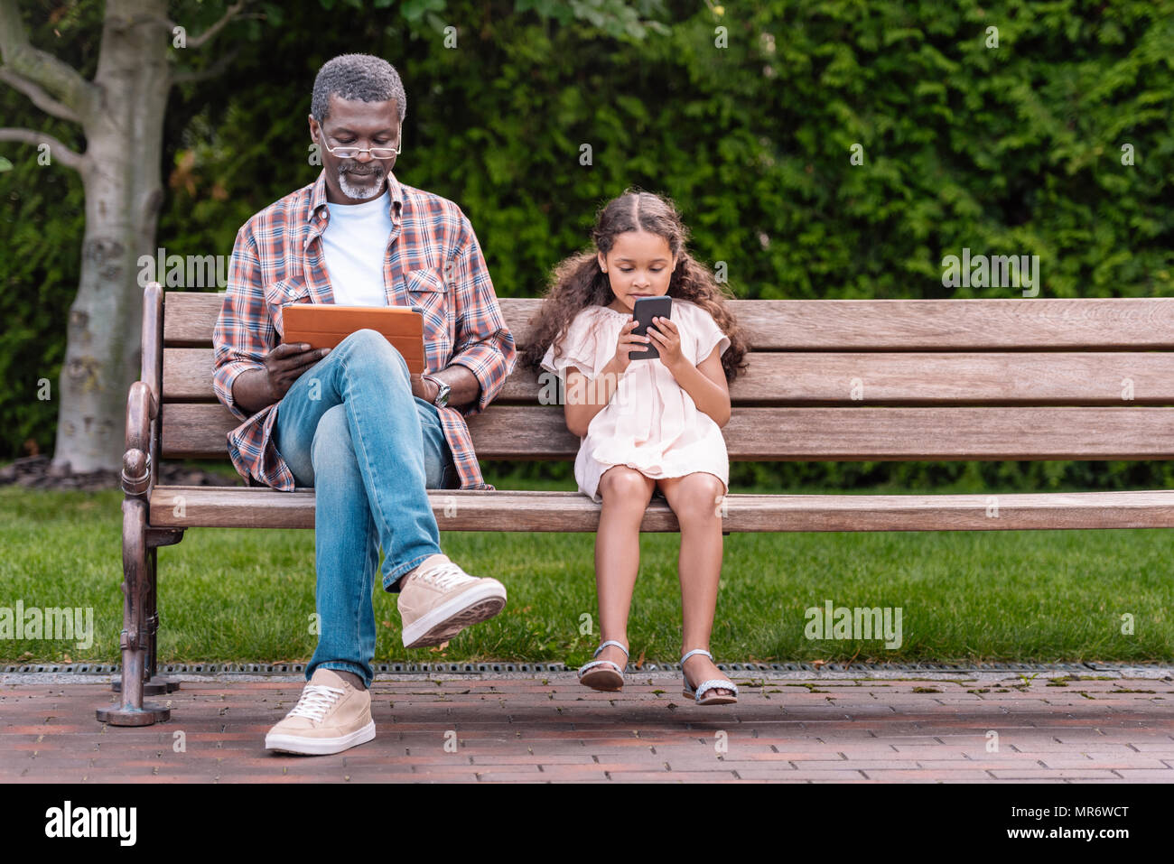 Adorable african american girl et son grand-père à l'aide de smartphone et tablette numérique tout en restant assis sur un banc dans le parc Banque D'Images