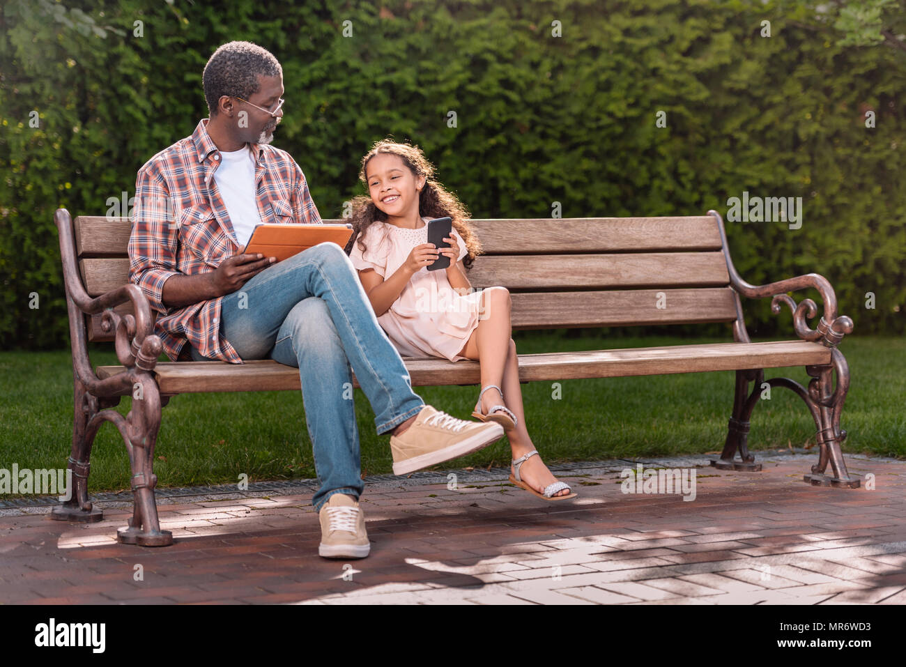 Smiling african american girl et son grand-père à l'aide de smartphone et tablette numérique tout en restant assis sur un banc dans le parc Banque D'Images
