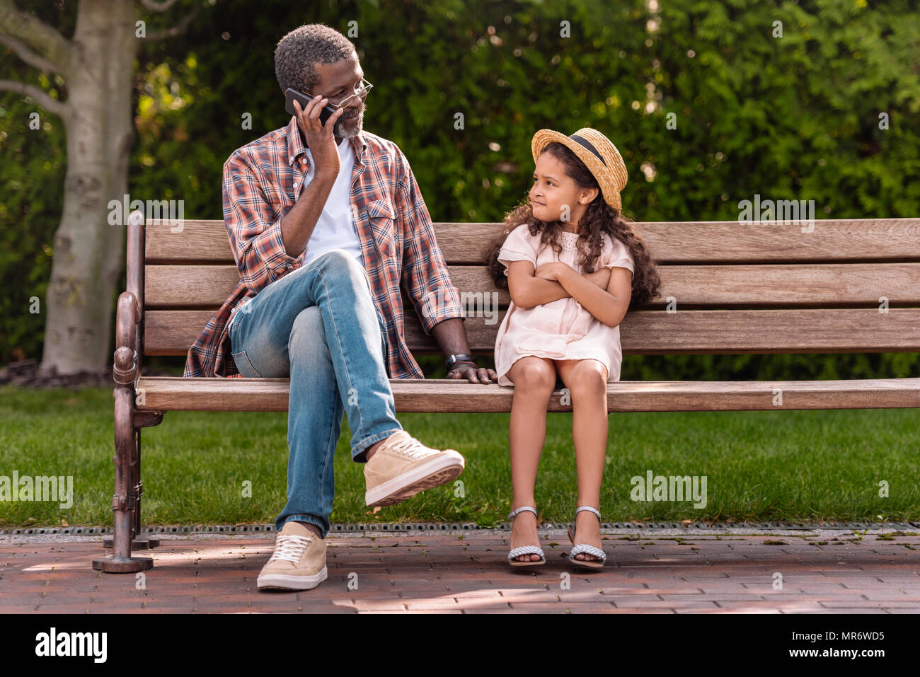 Petite-fille afro-américaine avec bras croisés à la recherche de son grand-père en conversation sur smartphone dans park Banque D'Images