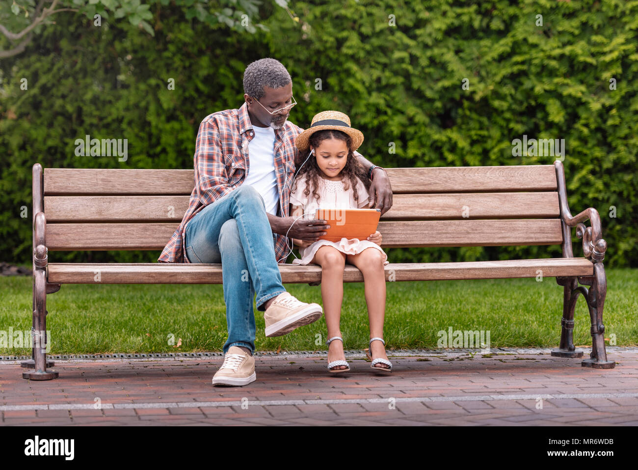Adorable petite-fille afro-américaine et de son grand-père à l'écoute de la musique sur digital tablet while sitting on bench in park Banque D'Images