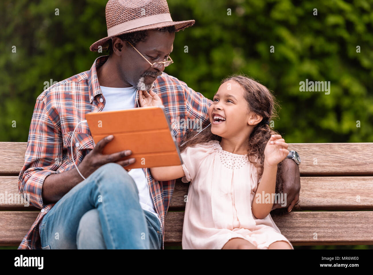 Happy african american petit-fils et son grand-père à l'écoute de la musique sur digital tablet while sitting on bench in park Banque D'Images