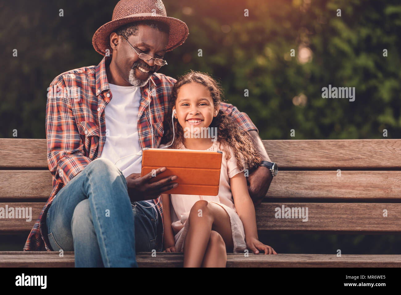 Smiling african american petit-fils et son grand-père à l'écoute de la musique sur digital tablet while sitting on bench in park Banque D'Images