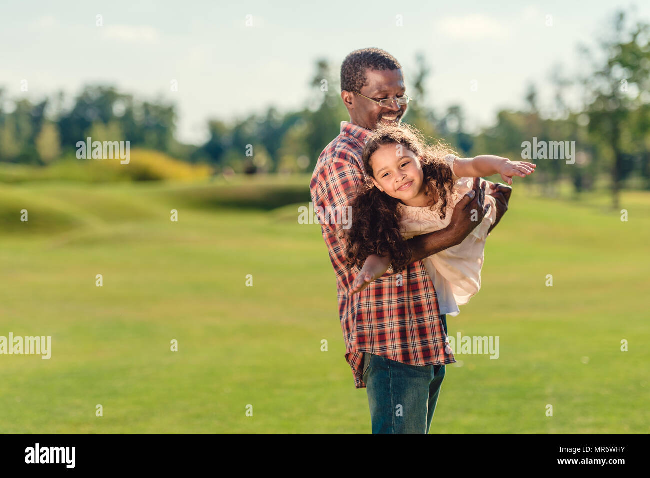 Smiling african american grand-père jouant avec plaisir sa petite-fille sur pelouse verte à jour ensoleillé Banque D'Images