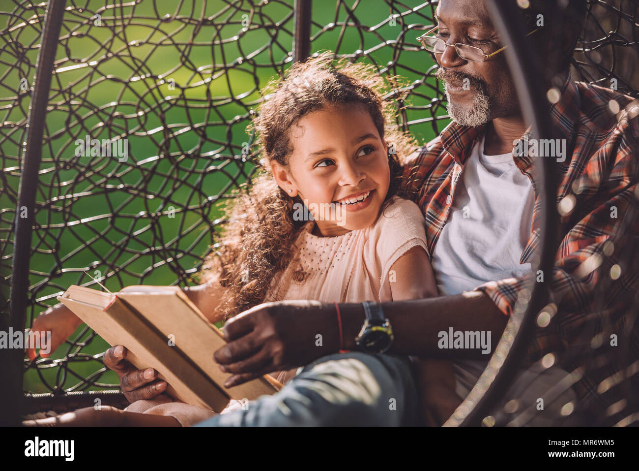 Smiling african american granddaughter reading book while sitting in swinging fauteuil suspendu avec son grand-père Banque D'Images