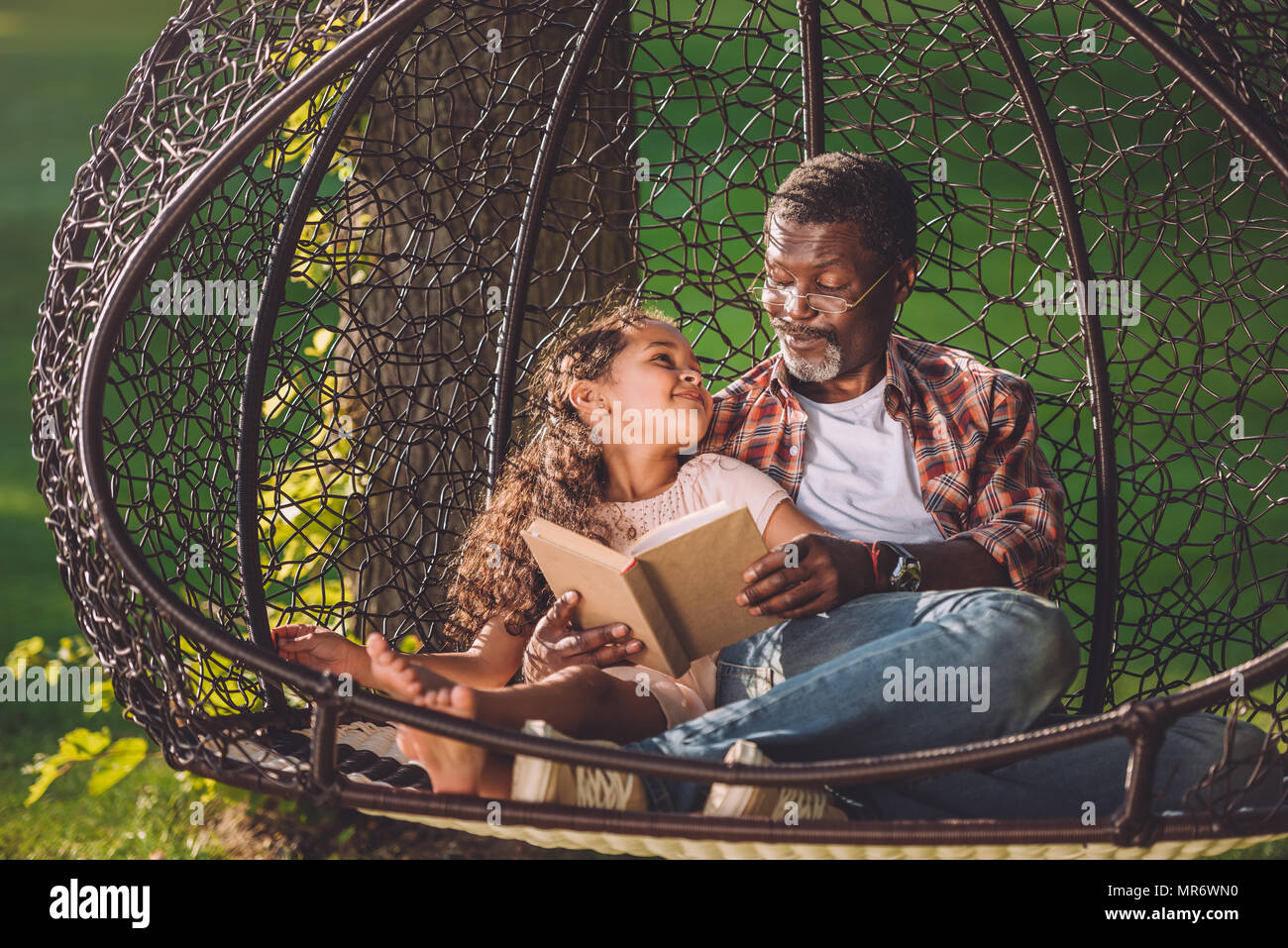 Happy african american petit-enfant livre de lecture tout en restant assis dans fauteuil suspendu oscillante avec grand-père Banque D'Images