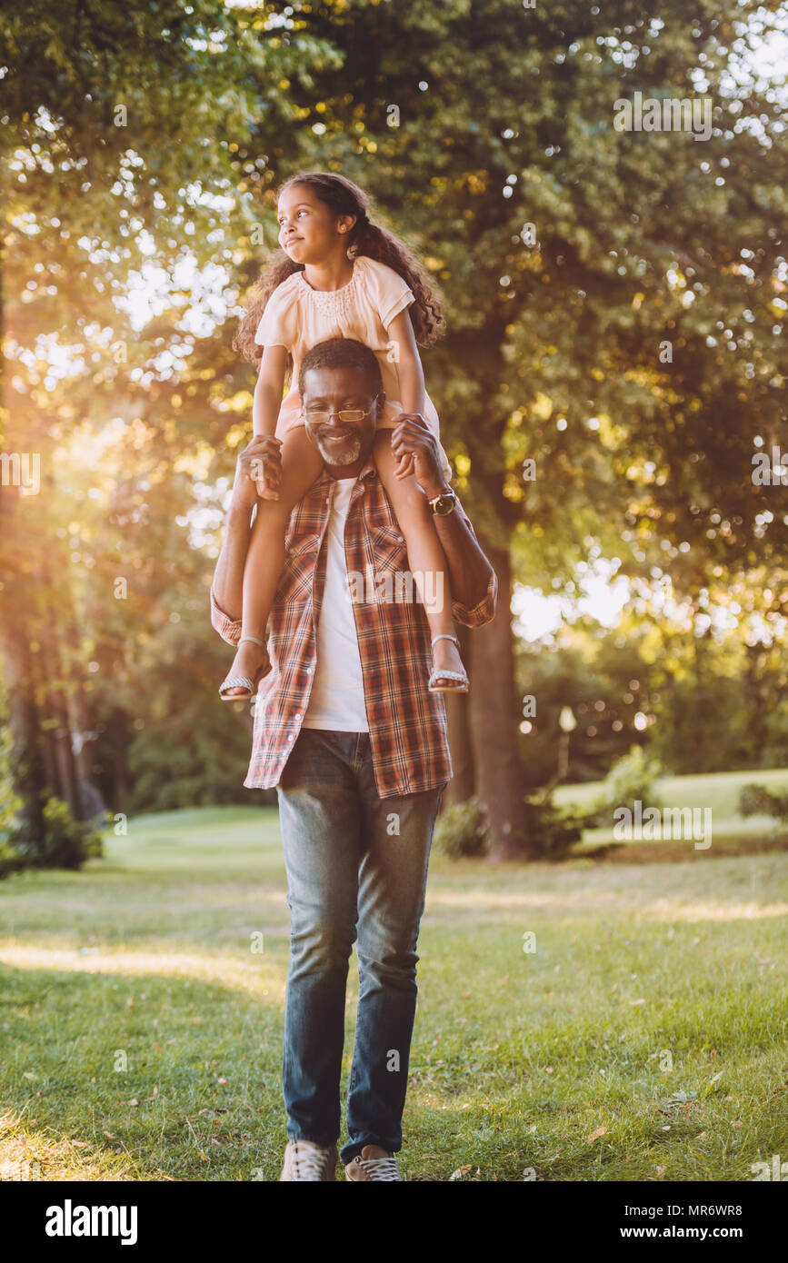 Happy african american petite fille assise sur les épaules de grand-père en parc avec lumière arrière Banque D'Images