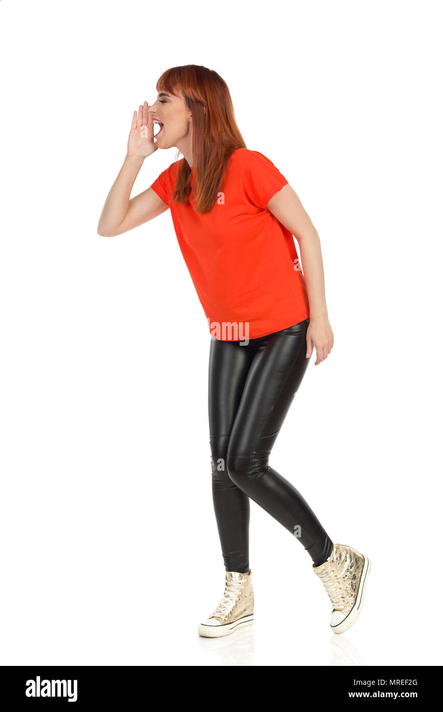 Jeune femme en tee-shirt orange, pantalon noir et or sneakers est debout, tenant la main sur le menton et crier. Vue de côté. Studio shot isolat pleine longueur Banque D'Images