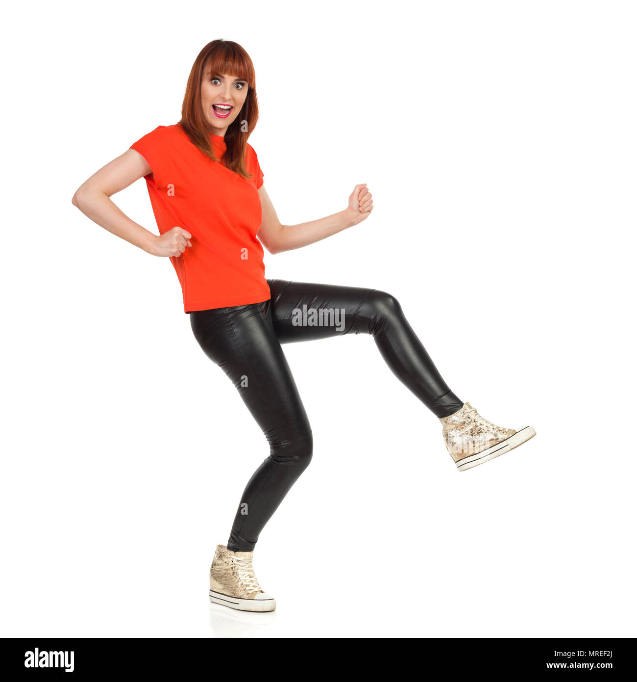 Jeune femme en tee-shirt orange, pantalon de cuir noir et or chaussures de marche est drôle et regardant la caméra. Vue de côté. Studio shot isolat pleine longueur Banque D'Images