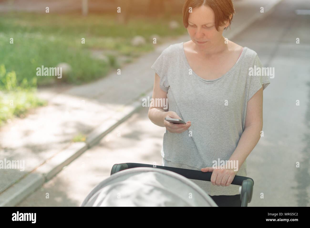 Mère poussant poussette de bébé et la lecture de texte message sur smart phone en marchant sur la rue en vêtements de sport décontracté Banque D'Images