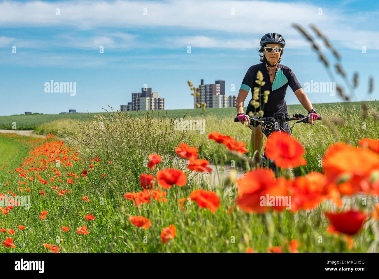 Nice senior woman riding a e-Mountainbike dans la banlieue d'une grande ville, entourée de champs verts et rouge coquelicot Banque D'Images