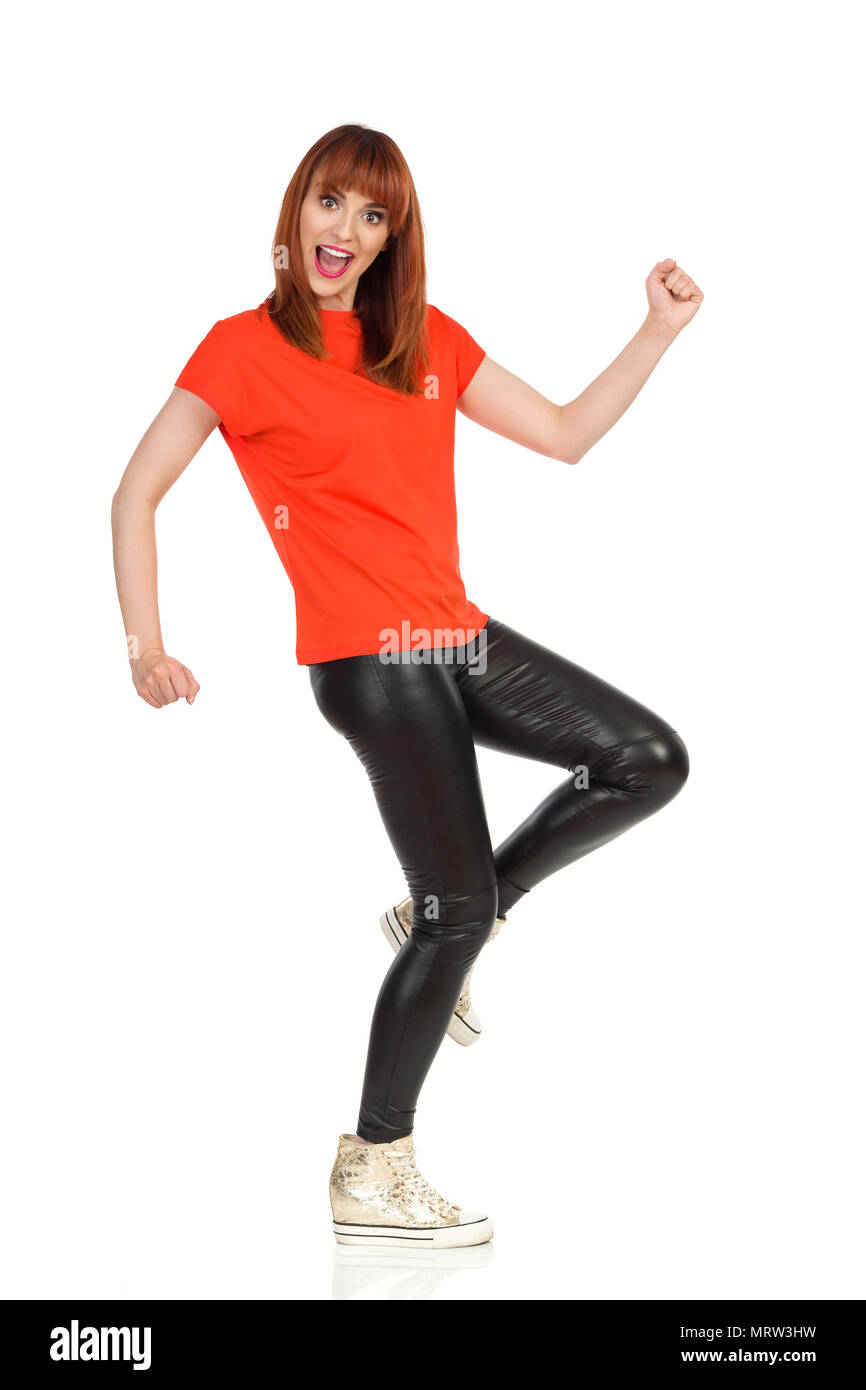 Jeune femme en tee-shirt orange, pantalon de cuir noir et or chaussures de marche est drôle, à la caméra et à parler. Vue de côté. Studio pleine longueur sh Banque D'Images