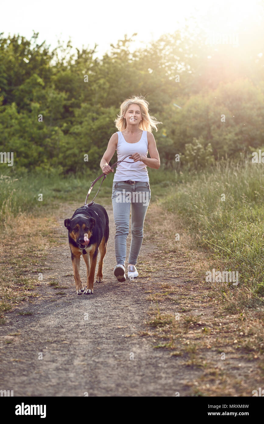 Happy middle-aged woman promener son chien le long d'une piste rurale d'herbe au printemps à la recherche de l'appareil photo Banque D'Images