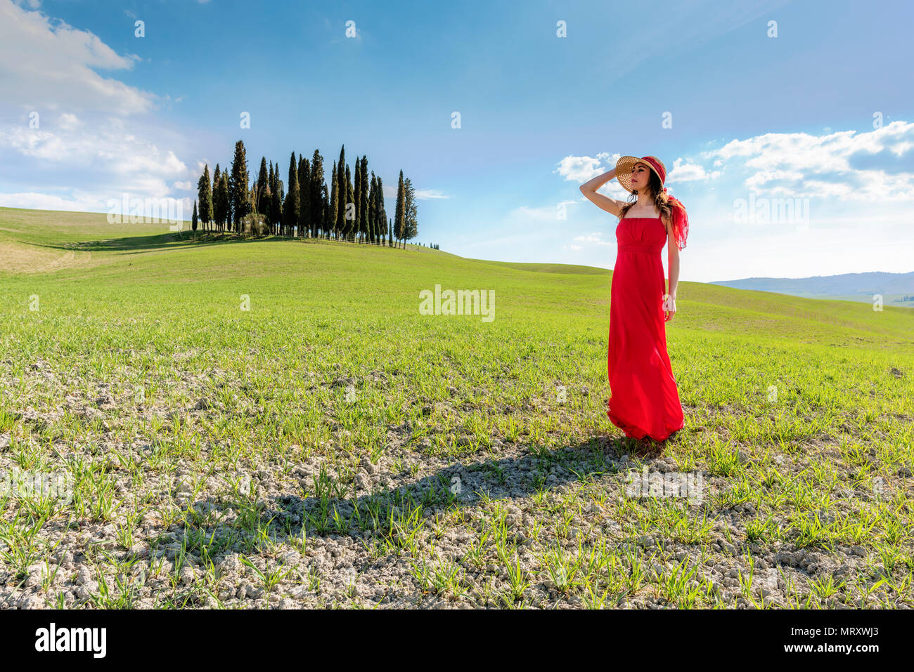 San Quirico d'Orcia, Val d'Orcia, Sienne, Toscane, Italie. Une jeune femme en robe rouge à admirer la vue dans un champ de blé près du cyprès de l'Orcia va Banque D'Images