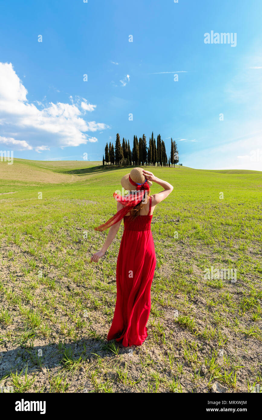 San Quirico d'Orcia, Val d'Orcia, Sienne, Toscane, Italie. Une jeune femme en robe rouge à admirer la vue dans un champ de blé près du cyprès de l'Orcia va Banque D'Images