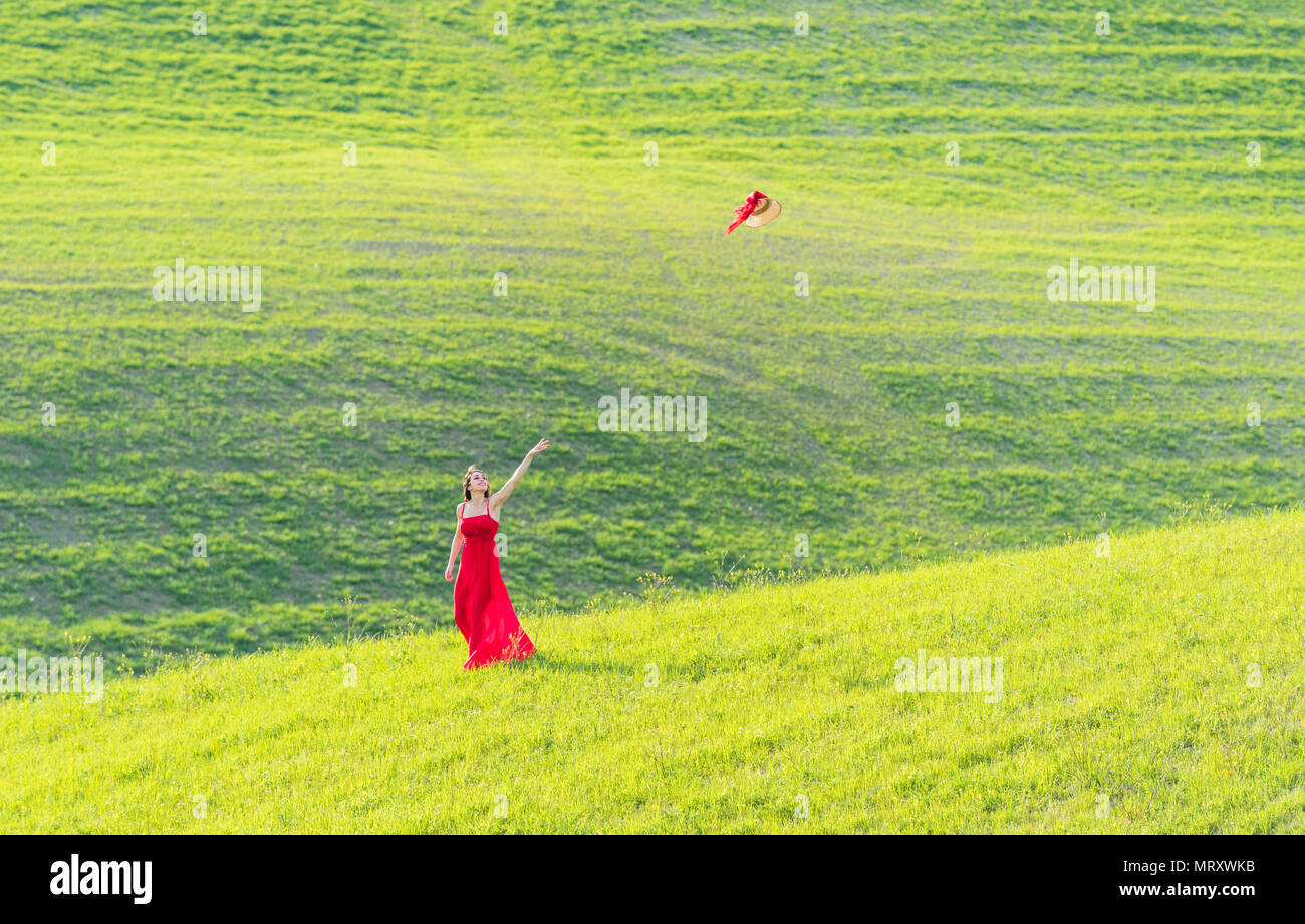 San Quirico d'Orcia, Val d'Orcia, Sienne, Toscane, Italie. Une jeune femme en robe rouge est de jeter son chapeau dans un champ de blé Banque D'Images