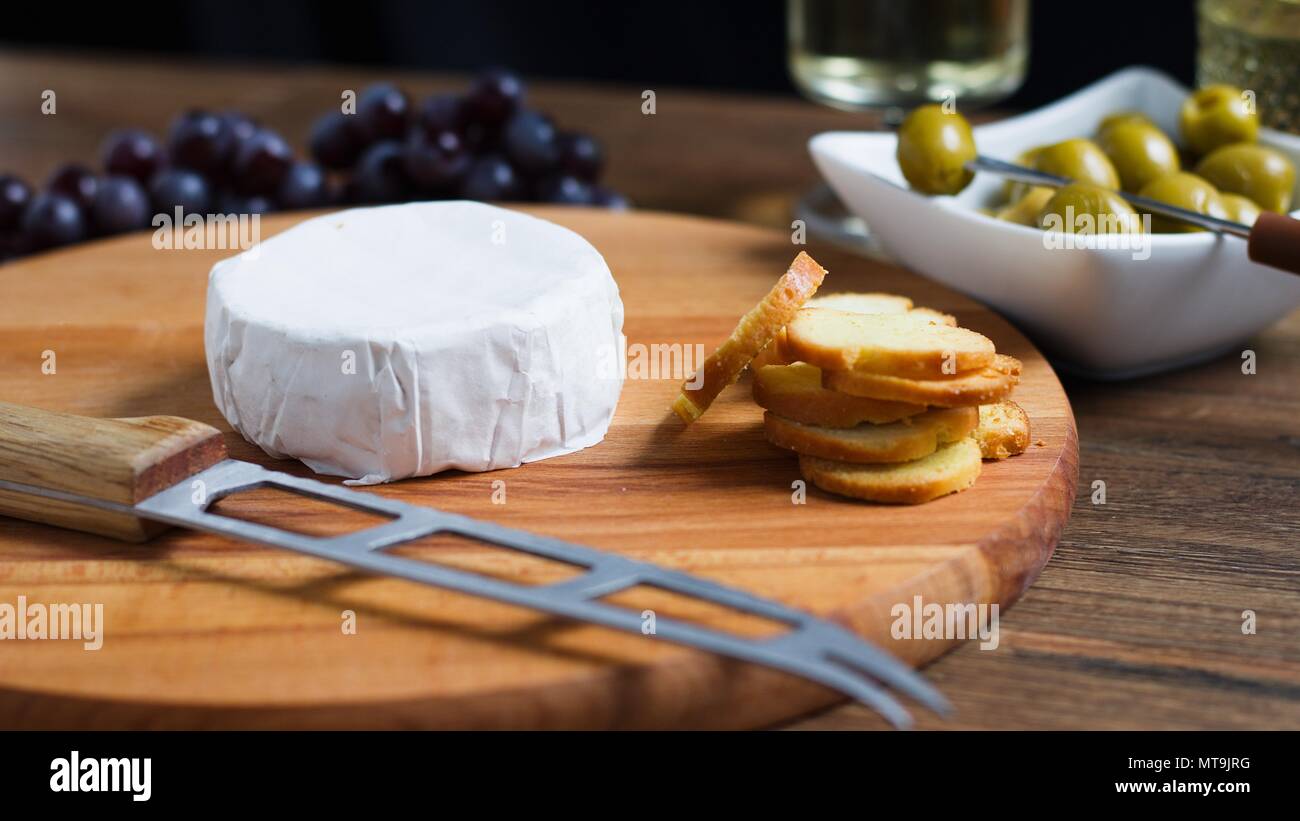 Fromage à pâte molle à croûte blanche (camembert ou brie) sur planche de bois avec des tranches de pain grillées, olives et vin blanc Banque D'Images