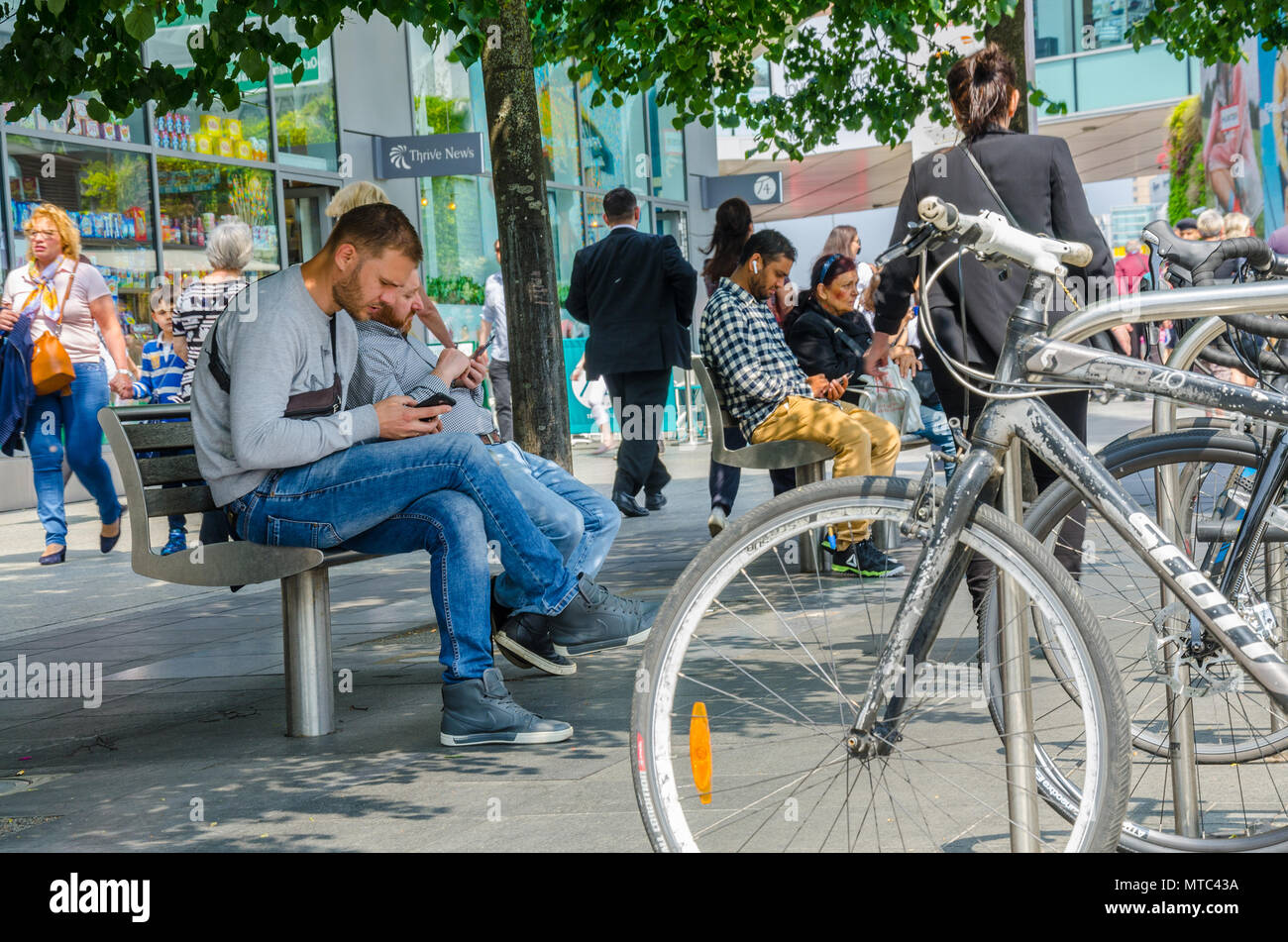 Les vélos garés dans un support à bicyclettes à côté de bancs avec les gens se préoccupe de leur téléphone mobile, Banque D'Images