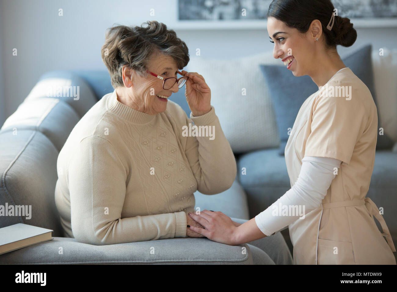 Une offre de soins de s'agenouiller et souriant face à une femme plus âgée qui est assis sur un canapé et tenant ses lunettes Banque D'Images