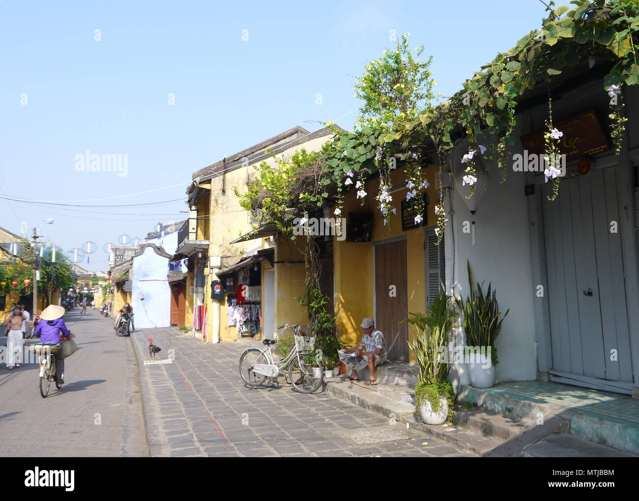 HOI AN, VIETNAM - 17ème Mars 2018 : Un homme assis sur un pas en avant d'un atelier fermé journal lecture début de matinée ensoleillée à Hoi An Banque D'Images