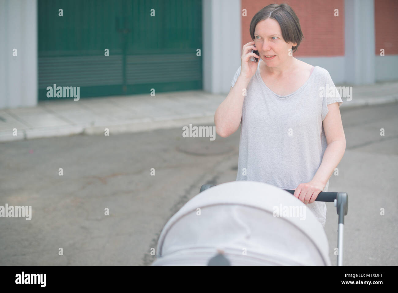 Contrarié et inquiet mother talking on mobile phone et en poussant bébé dans une poussette en marchant sur la rue en vêtements de sport décontracté Banque D'Images