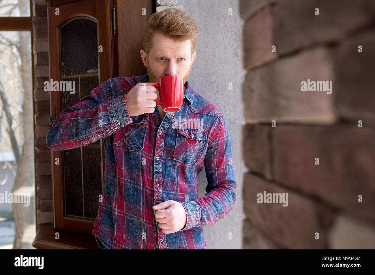 Homme avec neat hairstyle le café à l'extérieur de sa maison au petit matin. Cute gars avec golden ginger hair apprécie sa boisson chaude dans la tasse rouge vif leani Banque D'Images