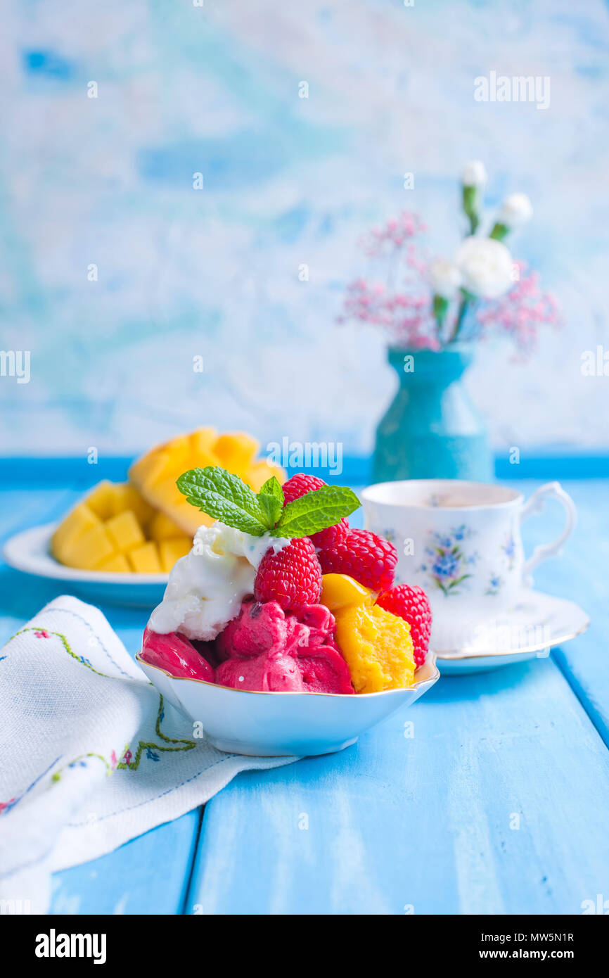 La crème glacée de fleurs aux couleurs vives avec des framboises et de la menthe dans une plaque, sur un fond en bois bleu. Une tasse de café le matin et de lumière et de fleurs. Vintage photo. Copy space Banque D'Images
