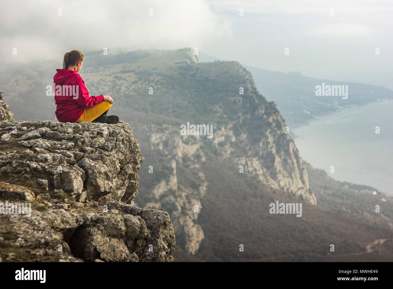Fille assise sur une falaise et à la recherche sur la mer et les montagnes avec des nuages Banque D'Images