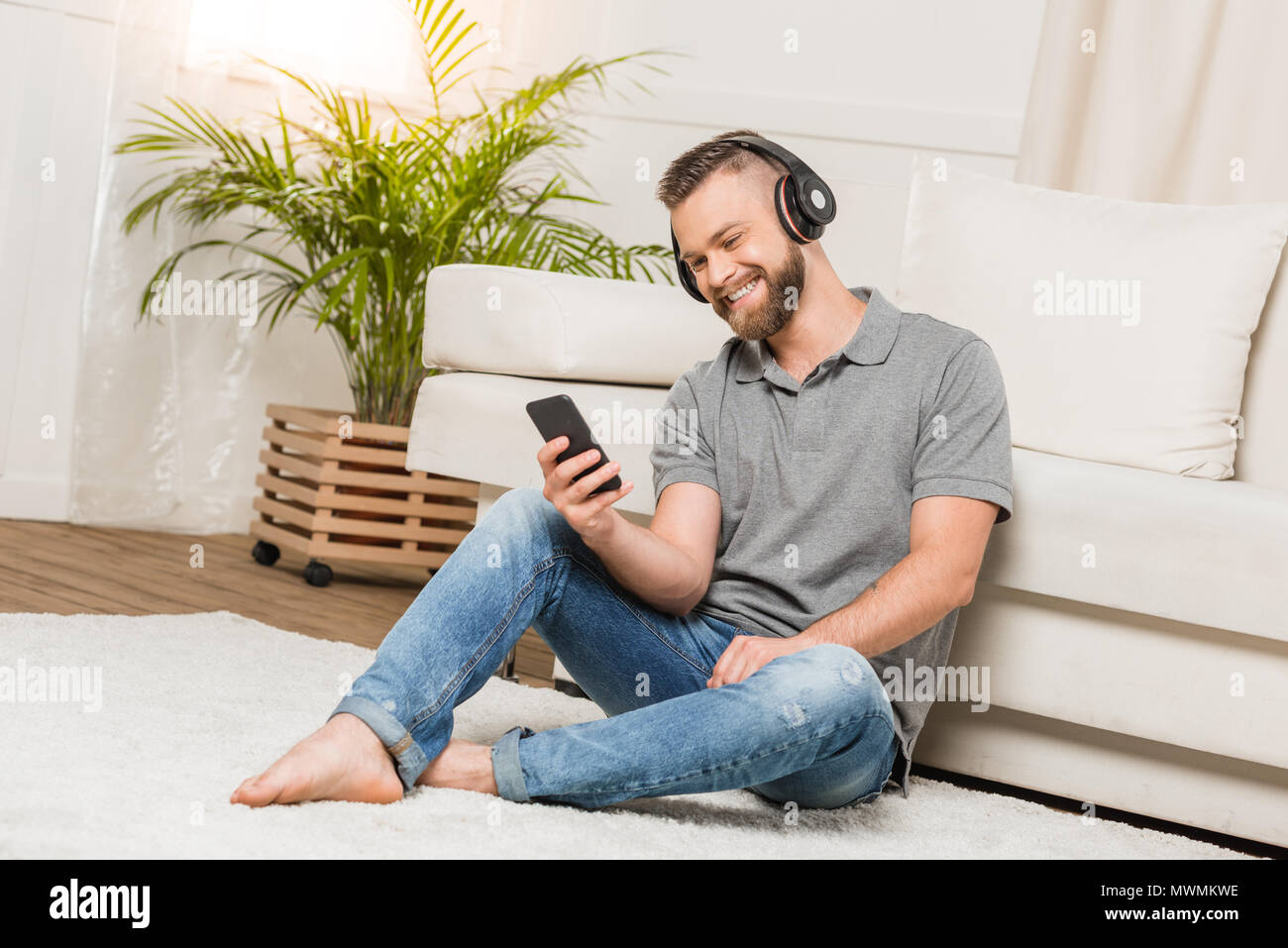 Jeune homme barbu joyeux dans les écouteurs sous smartphone et écouter la musique à la maison Banque D'Images