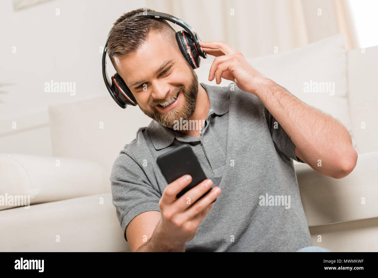 Jeune homme barbu joyeux dans les écouteurs sous smartphone et écouter la musique à la maison Banque D'Images