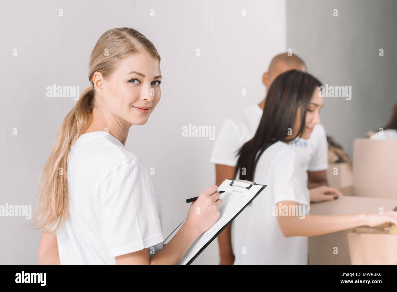 Les jeunes bénévoles de sexe féminin attrayant écrit dans le presse-papiers avec des collègues travaillant sur l'arrière-plan flou Banque D'Images