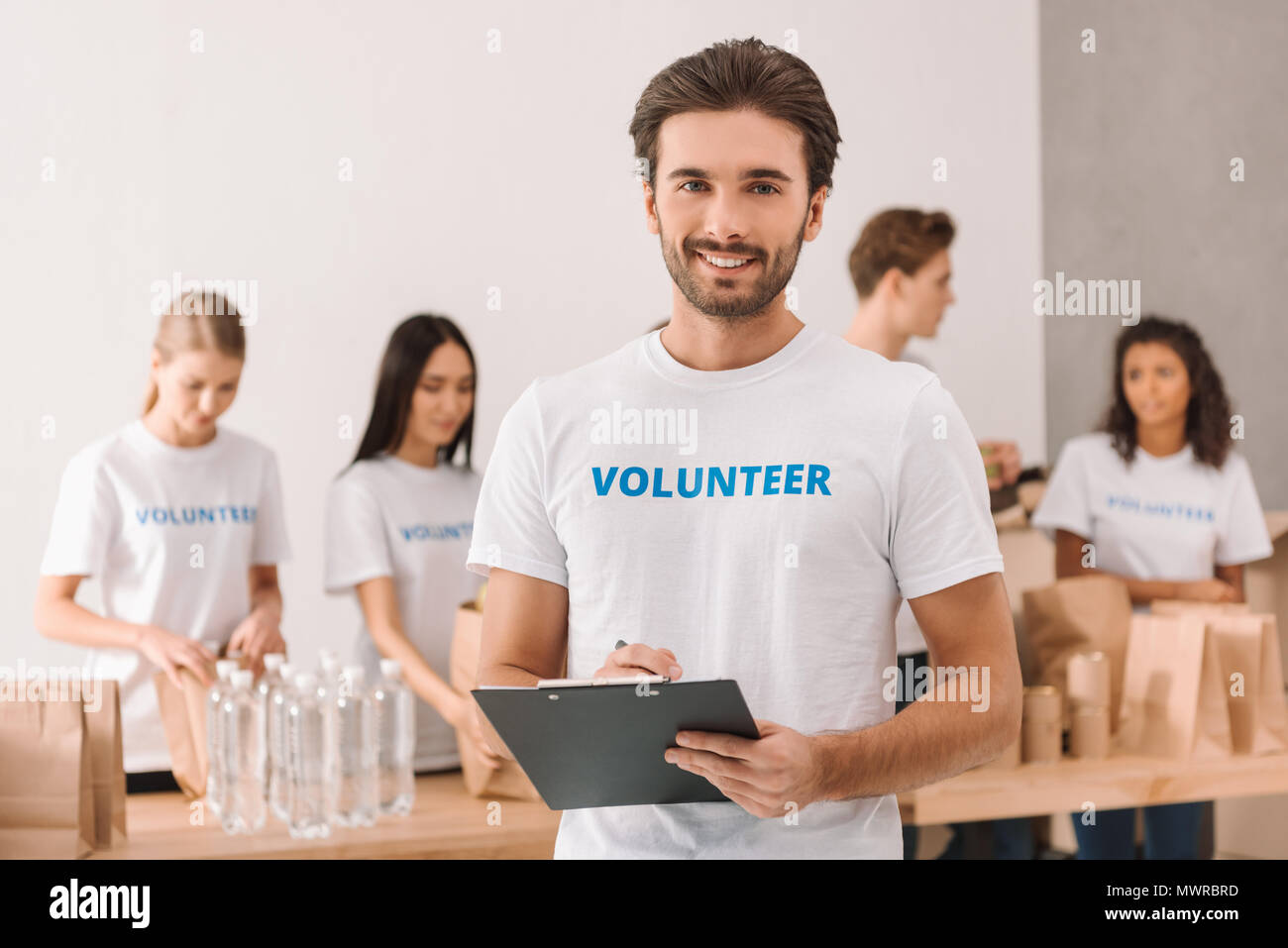 Belle écriture dans le presse-papiers bénévoles barbu et à la recherche à l'appareil photo avec des collègues travaillant sur l'arrière-plan flou Banque D'Images
