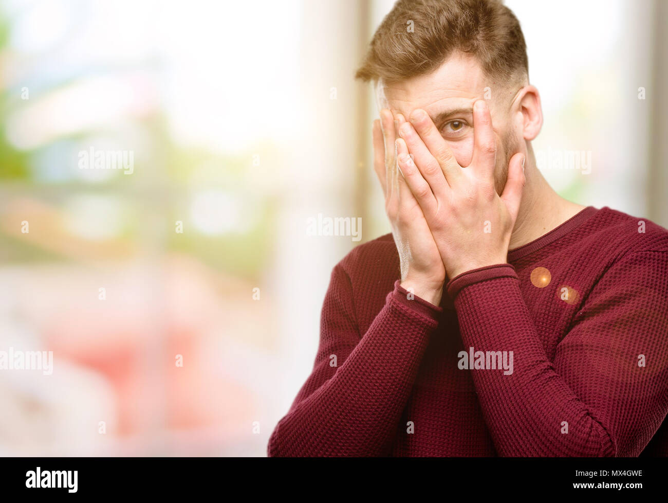 Beau jeune homme timide sourire avoir regarder furtivement à travers ses doigts, couvrant le visage avec les mains à l'ensemble confusément Banque D'Images