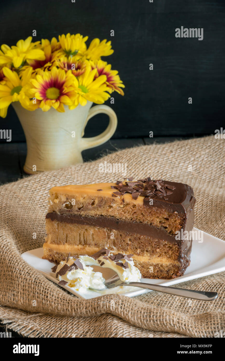 Tranche De Gateau Bresilien Avec Le Lait Et Le Chocolat De Remplissage Brigadeiro Servi Avec De La Creme Fouettee Sur Une Assiette Blanche Et Tissu Du Jute Et Fleurs Jaunes Photo Stock