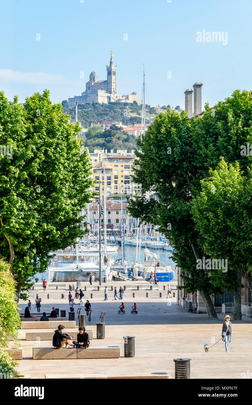 Les gens se promener ou se reposer sur place Villeneuve-Bargemon et derrière les arbres, le Vieux Port de Marseille et Notre-Dame de la Garde. Banque D'Images