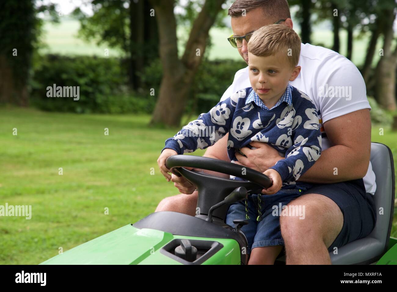 Père et fils : un père laisse son fils de 5 ans à diriger le tracteur-tondeuse qu'il coupe l'herbe Banque D'Images