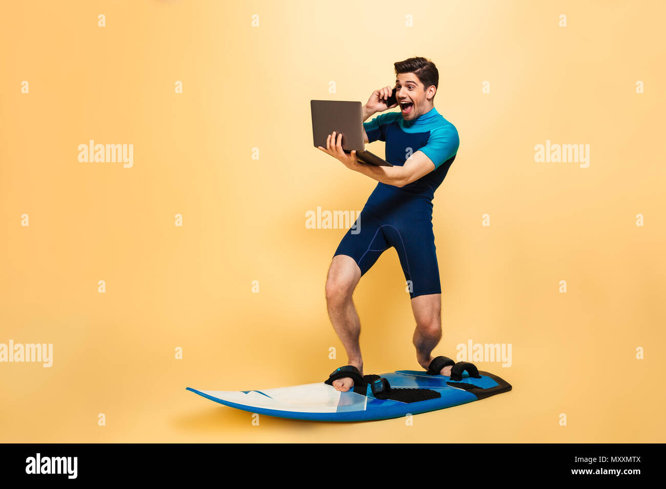 Photo d'un jeune homme excité habillés en maillot sur fond jaune isolé sur le surf board parler par téléphone à l'aide d'un ordinateur portable. Banque D'Images